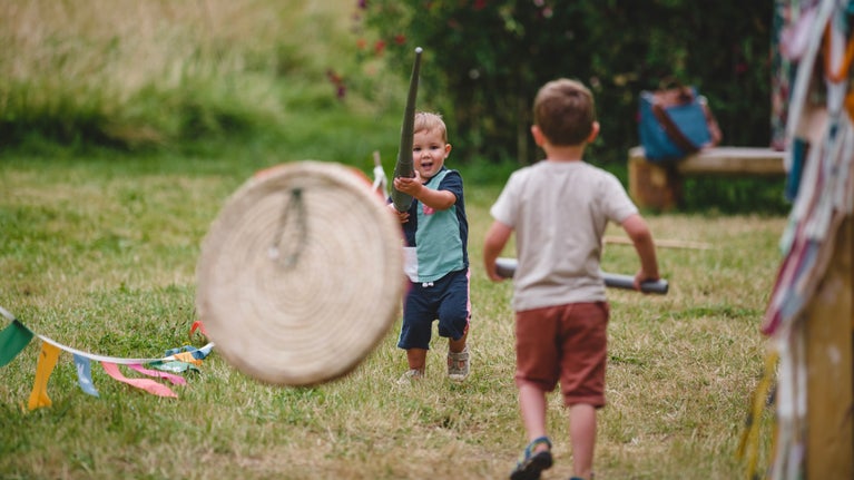 A young boy stands in the centre of the image with a toy lance. Another child is facing towards him, slightly out of focus, in the foreground, and colourful flags run alongside the children